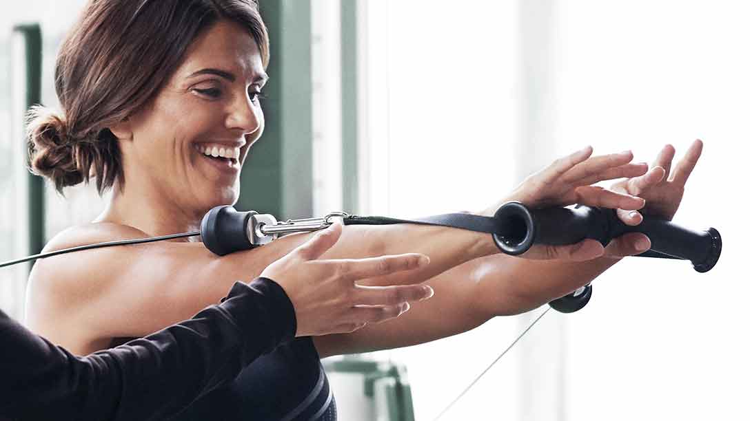 A smiling woman uses the dual cable cross machine with guidance from a trainer