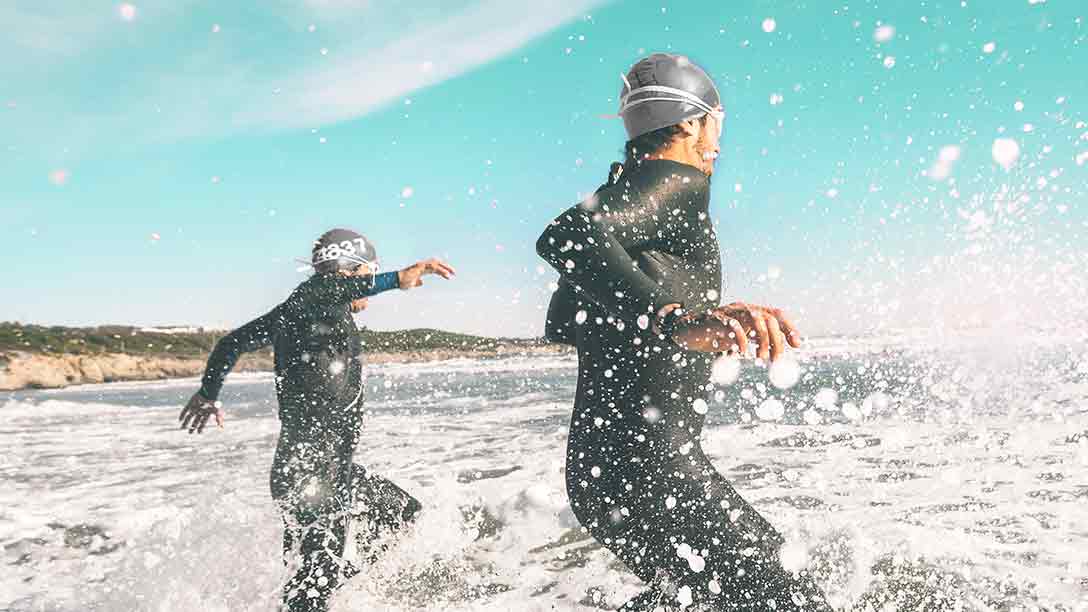 Two adults in swim caps and full-body wet-suits run into the ocean